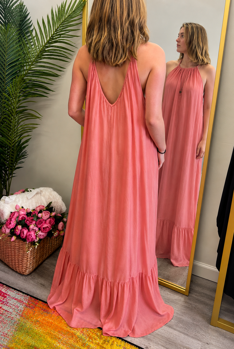 Woman in a pink dress standing in front of a mirror, with a plant and flowers in the background.