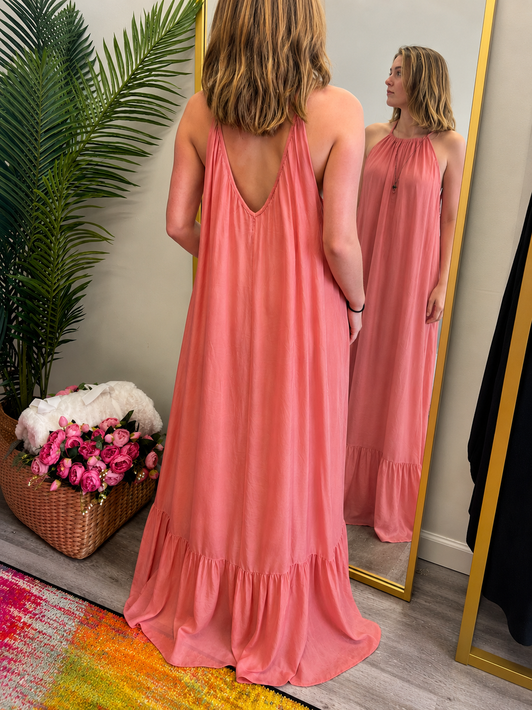 Woman in a pink dress standing in front of a mirror, with a plant and flowers in the background.